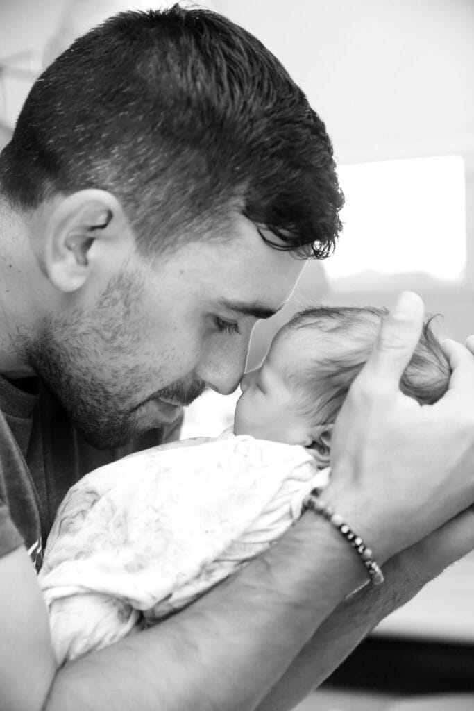 Man with beard pressing his forehead gently to a newborn baby he holds in both hands, black and white