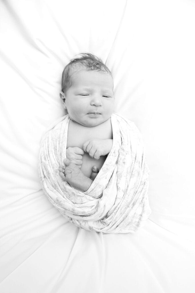 Newborn baby swaddled in a floral wrap lying on white fabric, black and white overhead view